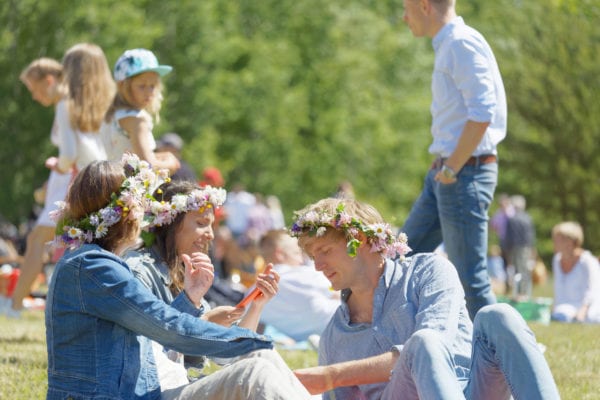 Two woman and a man with flower in their hair having a pic-nic sitting in the grass before celebrating the Midsummer in Sweden